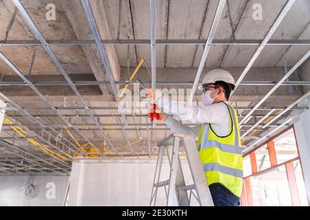 Asian handsome electrician wearing a mask installing laying electrical cables on the ceiling with pliers inside the house under construction. Stock Photo
