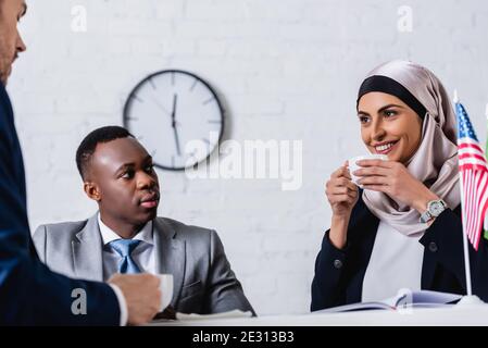 cheerful arabian businesswoman drinking coffee near multicultural business partners and american flag on blurred foreground Stock Photo