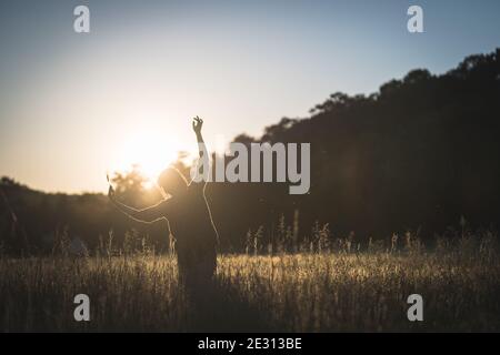 A young woman dances in the tall grass, backlit by the setting sun during golden hour Stock Photo