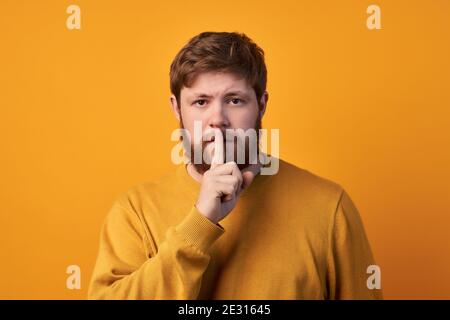 Photo of secret ginger stylish male guy shows silence sign, has surprised expression, afraids of revealing secret, dressed casually, poses alone again Stock Photo