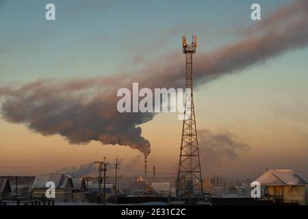 Smoke rises from the chimney of the boiler room over the city and the cell tower is in the foreground. Russia, Novosibirsk region, air temperature -41 Stock Photo