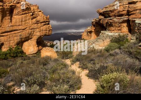 A small trail leading through a natural gateway between two large weathered sandstone boulders in the Cederberg Mountains of South Africa, with a dark Stock Photo