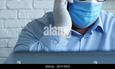 Doctor in a Hospital Office with a Face Mask and Protective Gloves Use Smartphone in Front of the Laptop Screen Stock Photo