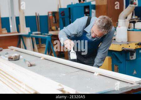 An elderly cabinetmaker in overalls and glasses paints a wooden board with a brush on a workbench in a carpentry shop.  Stock Photo