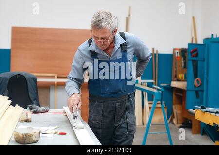 An elderly cabinetmaker in overalls and glasses paints a wooden board with a roller on a workbench in a carpentry shop. Stock Photo