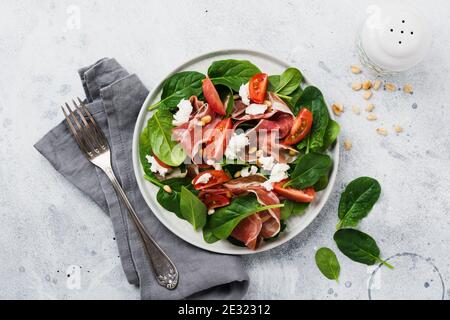 Spring Salad with spinach, cherry tomato, mozzarella, pine nuts and ham with olive oil in a simple ceramic plate on an old concrete gray background. Stock Photo