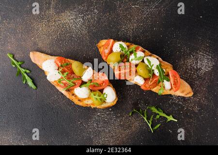 Caprese bruschetta toasts with cherry tomatoes, mozzarella, olives and basil on old dark background. Top view. Stock Photo