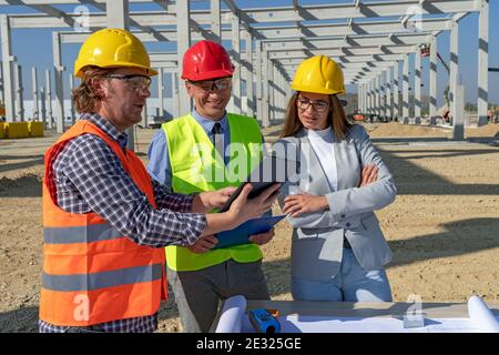 Construction Foreman in Yellow Hardhat Holding Digital Tablet and Discuss a Project With Female Architect and Businessman. Teamwork Concept. Stock Photo