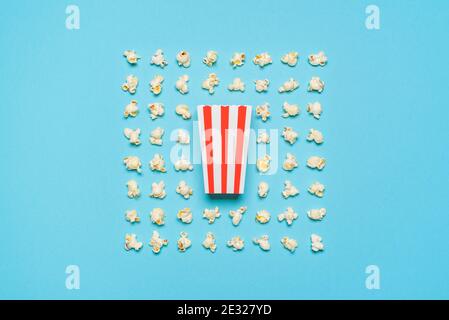 Above view with a striped cardboard box and many pop-corn flakes aligned symmetrically on a blue-colored background. Pop-corn box isolated on a seamle Stock Photo