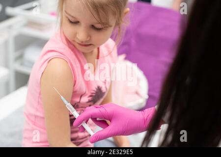 The nurse gently inserts the needle into the little girl's arm. The child looks at the puncture site with the needle. A doctor's office in a public hospital. Safe vaccination of children. Stock Photo