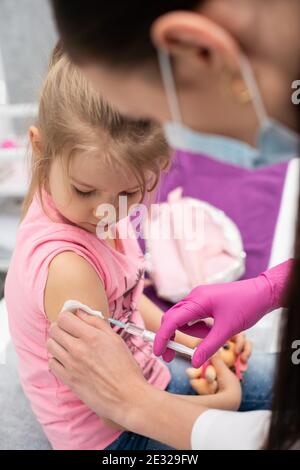 The nurse gently inserts the needle into the little girl's arm. The child looks at the puncture site with the needle. A doctor's office in a public hospital. Safe vaccination of children. Stock Photo