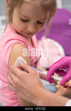 The nurse gently inserts the needle into the little girl's arm. The child looks at the puncture site with the needle. A doctor's office in a public hospital. Safe vaccination of children. Stock Photo