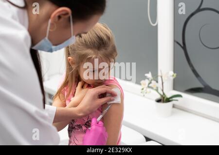 The nurse gently inserts the needle into the little girl's arm. The child looks at the puncture site with the needle. A doctor's office in a public hospital. Safe vaccination of children. Stock Photo