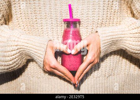 Woman in woollen sweater making heart shape with her hands and holding bottle with pink smoothie or juice Stock Photo