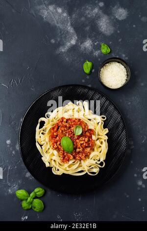 Traditional Italian dish fettuccine pasta with bolognese sauce, basil and parmesan cheese in black plate on dark wooden background. Top view. Stock Photo