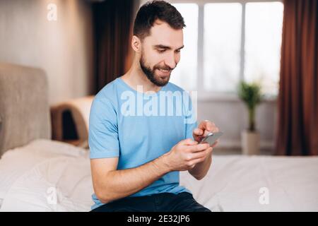 Young man with beard smiling whilst received sending message on phone while sitting in bed. Stock Photo