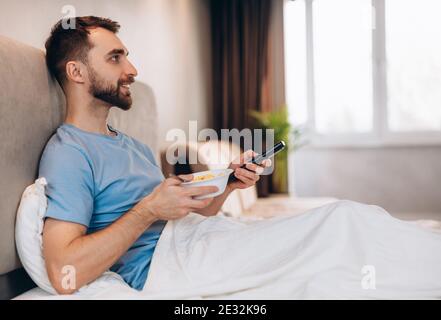 Handsome young man with beard having breakfast in bed and watching tv. weekend lazy day off. Stock Photo