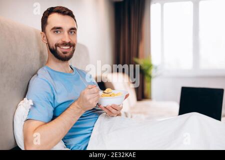 Handsome young man with beard smiling eating breakfast in bed look at camera. weekend lazy day off. Stock Photo