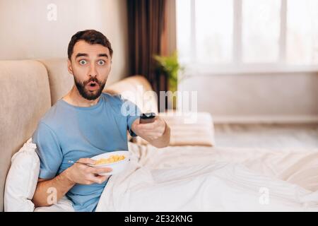 Attractive young man with beard eating breakfast in bed and look disappointed shocked whilst watching news. Stock Photo
