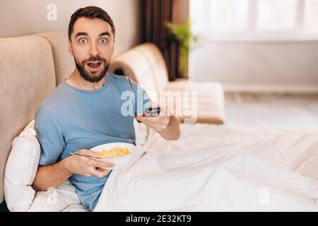Attractive young man with beard eating breakfast in bed and look surprised whilst watching news. Stock Photo