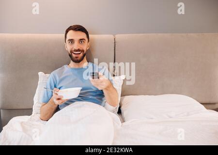 Attractive young man with beard eating breakfast in bed and look happy whilst watching tv shows. Stock Photo