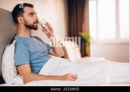 Handsome young man with beard smiling drinking water before breakfast in bed look at camera. weekend lazy day off. Stock Photo