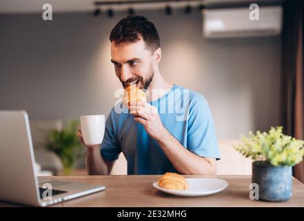 Daily life of young man in kitchen during breakfast using laptop holding a cup of coffee. Attractive male working from home, telecommuting using Stock Photo