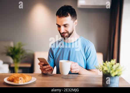 Daily life of young man in kitchen during breakfast using smartphone holding a cup of coffee. Attractive male working from home, telecommuting using Stock Photo