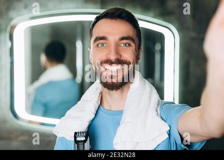 Young smiling man with towel holding brand-new shaver and taking a selfie early in the morning at bathroom. Stock Photo