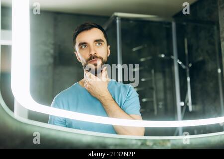 attractive young man with blue eyes touching his beard in front of mirror in the morning. Stock Photo