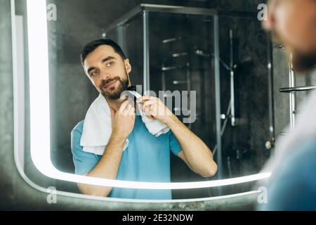 Young handsome man with towel looking at mirror and shaving early in the morning at home. Stock Photo