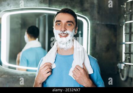 Handsome young man standing with towel on neck and foam on face smiling while looking at camera. Stock Photo