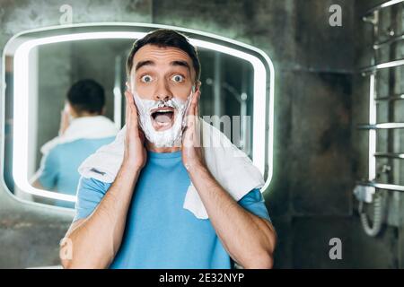 Handsome young man standing with towel on neck and foam on face while looking scared surprised at camera. Stock Photo