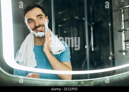 Close-up shot of smiling young man with towel in his neck standing in front of mirror and applying shaving foam to his face. Stock Photo