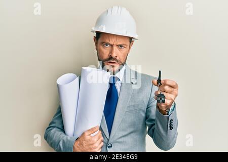 Middle age architect man wearing safety helmet holding blueprints and new home key in shock face, looking skeptical and sarcastic, surprised with open Stock Photo