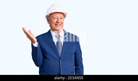 Senior handsome grey-haired man wearing suit and architect hardhat smiling cheerful presenting and pointing with palm of hand looking at the camera. Stock Photo