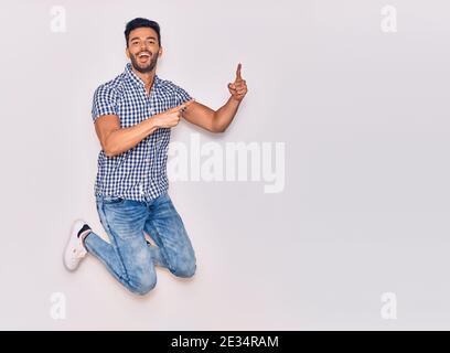 Young handsome hispanic man wearing casual clothes smiling happy. Jumping with smile on face pointing with fingers to the side over isolated white bac Stock Photo