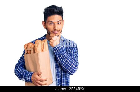 Handsome latin american young man holding paper bag with bread looking confident at the camera smiling with crossed arms and hand raised on chin. thin Stock Photo