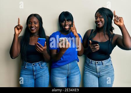 Three young african american friends using smartphone smiling amazed and surprised and pointing up with fingers and raised arms. Stock Photo
