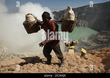 Miner Pak Agus, the oldest sulphur miner, carries the sacks with sulphur along the edge of the crater of the active volcano of Kawah Ijen in East Java, Indonesia. Pak Agus is 60 year old now. He works at the sulphur mines located down in the crater for more than 40 years. When he was younger, he was able to carry more than 100 kilograms of sulphur up from the bottom of the crater. Now his limit is 80 kilograms. Pak Agus is the only working miner of his age and he is quite lucky to be still alive. Most of miners die at age around 40. The acid lake at the bottom of the crater, one of the largest Stock Photo