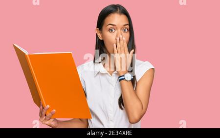 Beautiful hispanic woman reading book covering mouth with hand, shocked and afraid for mistake. surprised expression Stock Photo