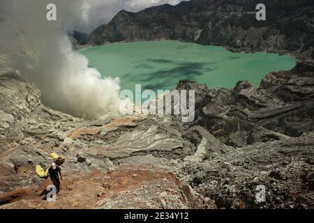Miners lift the baskets with sulphur from the sulphur mines located in the crater of the active volcano of Kawah Ijen in East Java, Indonesia. The acid lake at the bottom of the crater, one of the largest volcanic lakes in the world, is seen in the background. The lake is filled with a poisonous mix of sulphuric and hydrochloric acids; thanks to this it has its milky turquoise colour. Every morning two hundreds miners climb down to the bottom of the crater for the next load of sulphur. They fill their baskets with sulphur manually and then carry that heavy load up on feet. Extremely hard work Stock Photo