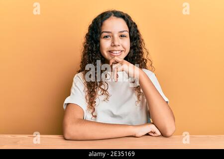 Teenager hispanic girl wearing casual clothes sitting on the table looking confident at the camera with smile with crossed arms and hand raised on chi Stock Photo