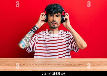 Young hispanic man using headphones sitting on the table in shock face, looking skeptical and sarcastic, surprised with open mouth Stock Photo