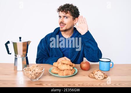 Young caucasian man with curly hair sitting on the table having breakfast smiling with hand over ear listening and hearing to rumor or gossip. deafnes Stock Photo
