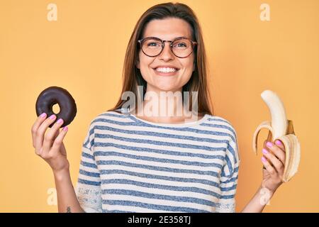Beautiful caucasian woman holding banana and chocolate doughnut smiling with a happy and cool smile on face. showing teeth. Stock Photo