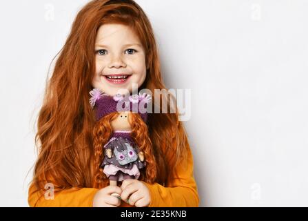 Cute happy smiling red-haired kid girl in orange sweatshirt holds small redhair doll her portrait in hands at face Stock Photo
