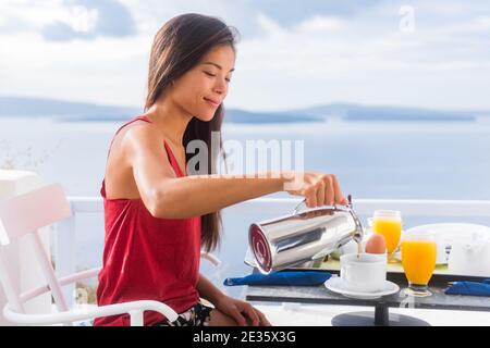 Coffee woman pouring tea in mug for breakfast at hotel room by Mediterranean sea on Europe vacation travel. Happy Asian girl enjoying morning brunch