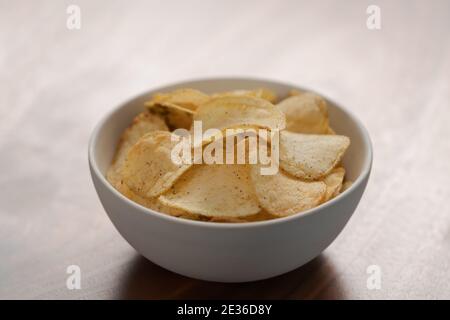 White bowl with organic potato chips sprinkled with fresh ground black pepper on wood background, shallow focus Stock Photo