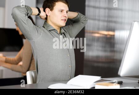 Young businessman and programmer with brown curly hair in a green shirt is stretching his hands after working hard on computer, sitting together with Stock Photo
